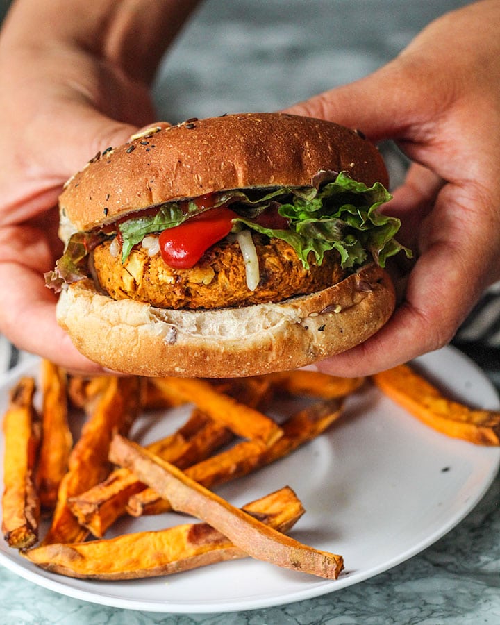 Chickpea burger assembled and being held. Burger is topped with vegan cheese, ketchup and lettuce between two buns and served with sweet potato fries.
