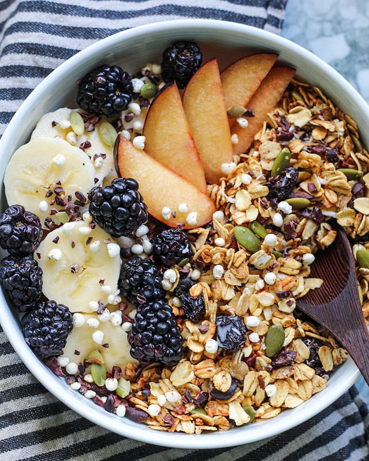 Fresh bowl of granola with milk topped with sliced plums, banana and blackberries.