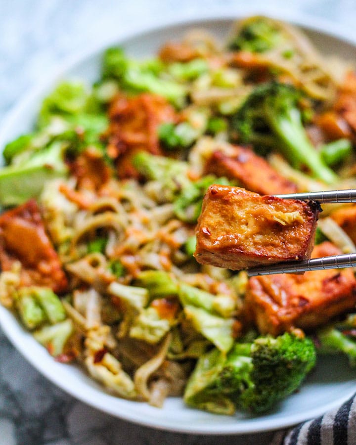 A close up of the garlic chili tofu coated with peanut sauce and held up with a chopstick.