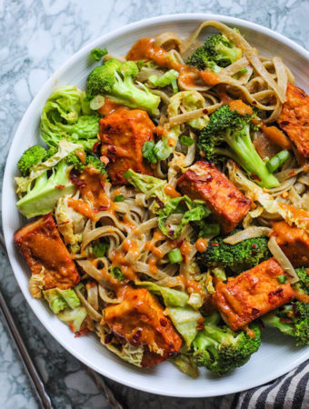 Bowl of baked garlic chili tofu mixed with broccoli, cabbage and rice noodles then tossed together with peanut sauce.
