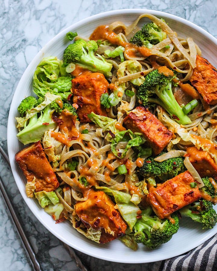 Bowl of baked garlic chili tofu mixed with broccoli, cabbage and rice noodles then tossed together with peanut sauce.