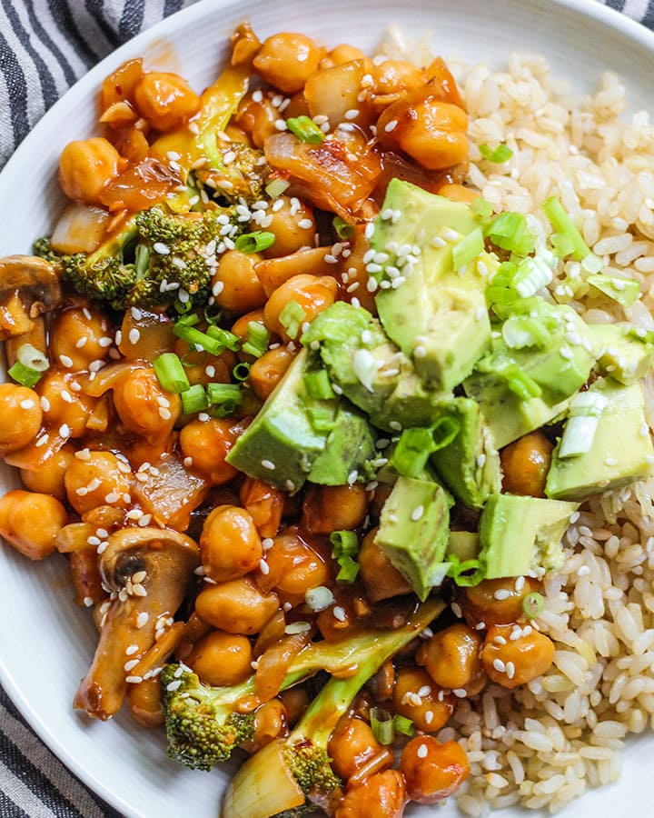 Plate of rice, sweet and sour chickpeas, avocado and topped with diced spring onion and sesame seeds.