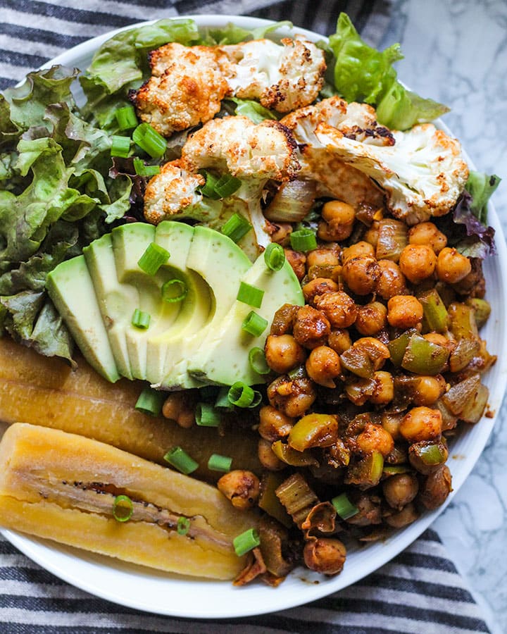 Seasoned chickpeas served on a plate with boiled plantains, greens, avocado and roasted cauliflower.