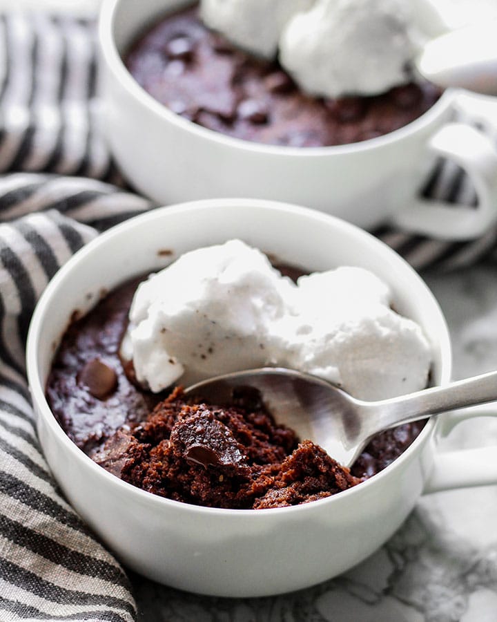 Side angle shot of two vegan brownie mugs topped with ice cream with a spoonful of it upfront.