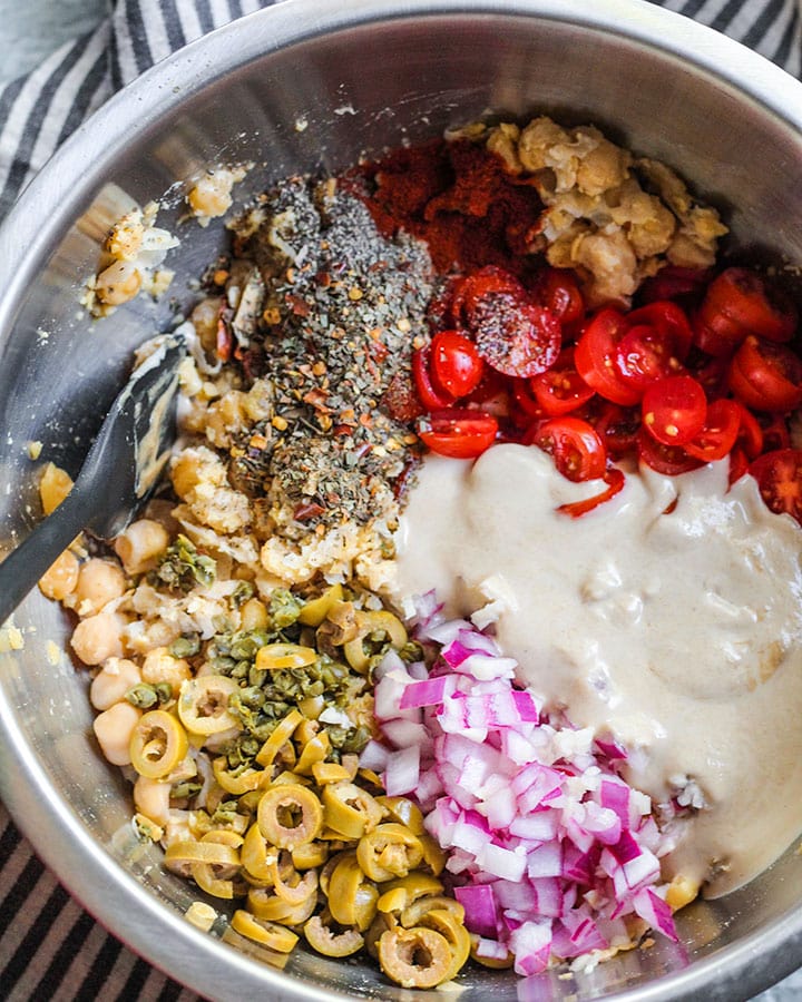 A mixing bowl with mashed chickpeas, minced onion and garlic, sliced green olives, papirka, oregano, pepper, basil, tahini and lemon.