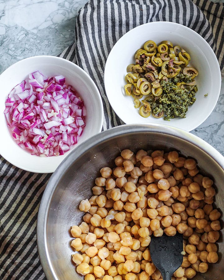 A bowl of chickpeas, a bowl of onion and garlic, and a bowl of sliced green olives and minced capers.