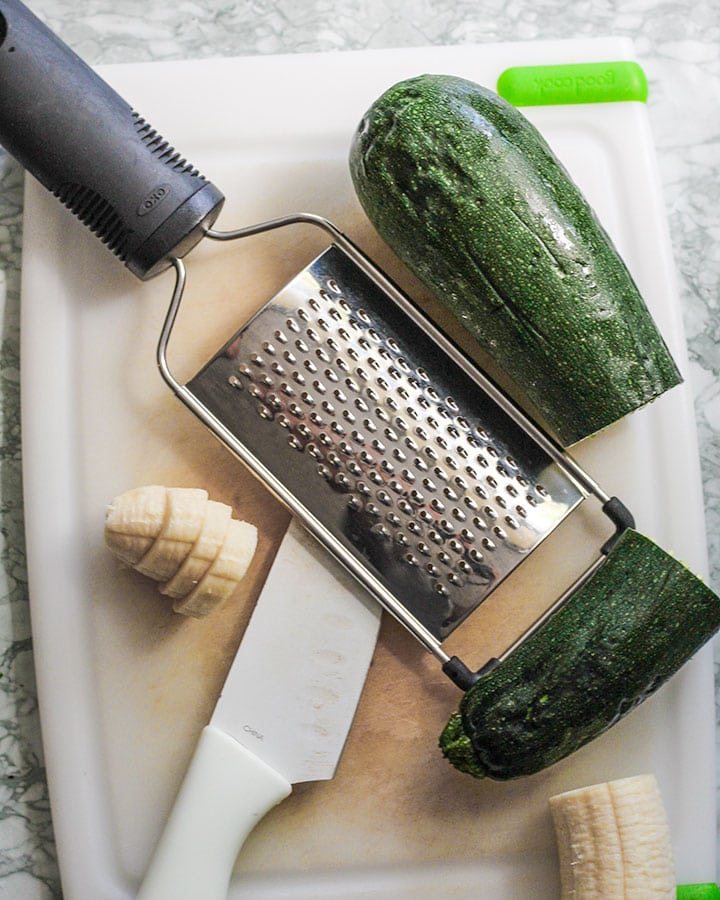 Prepping a large zucchini to grate using a grater and prepping banana to garnish with oats.