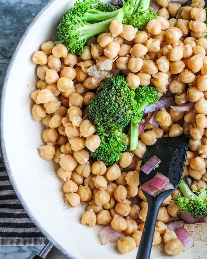 Sauted chickpeas, red onion, broccoli, and mushrooms in a pan with spatula.