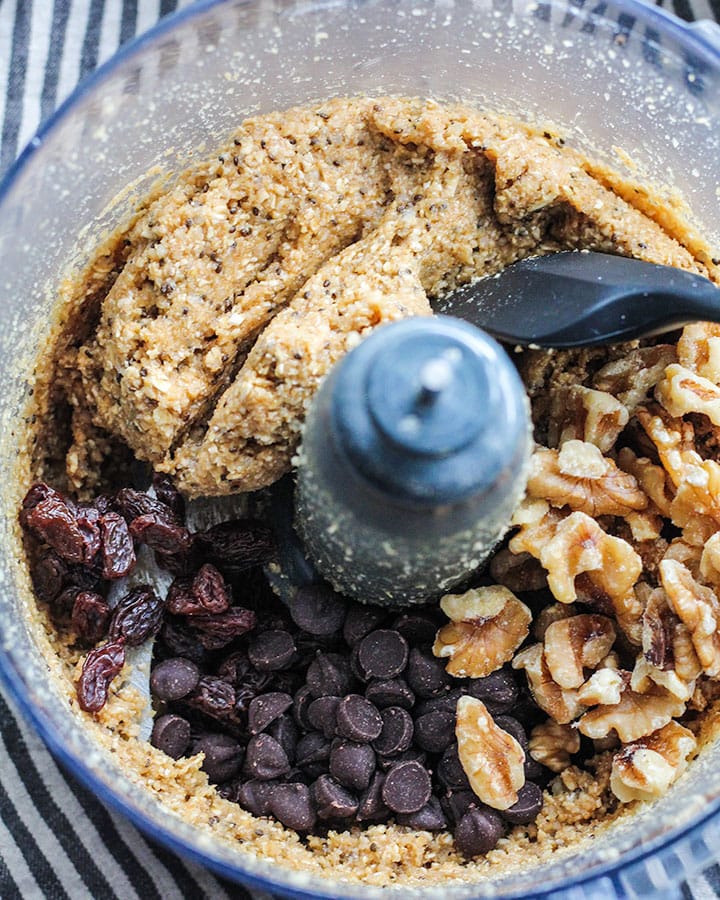 Formed cookie dough in the food processor now topped with cookie mix in ingredients including chocolate chips, walnuts and raisins. 