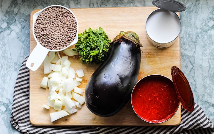 Cutting board with a cup of lentils, diced onion, diced kale, one whole eggplant, one can of crushed tomatoes and one can of coconut milk.