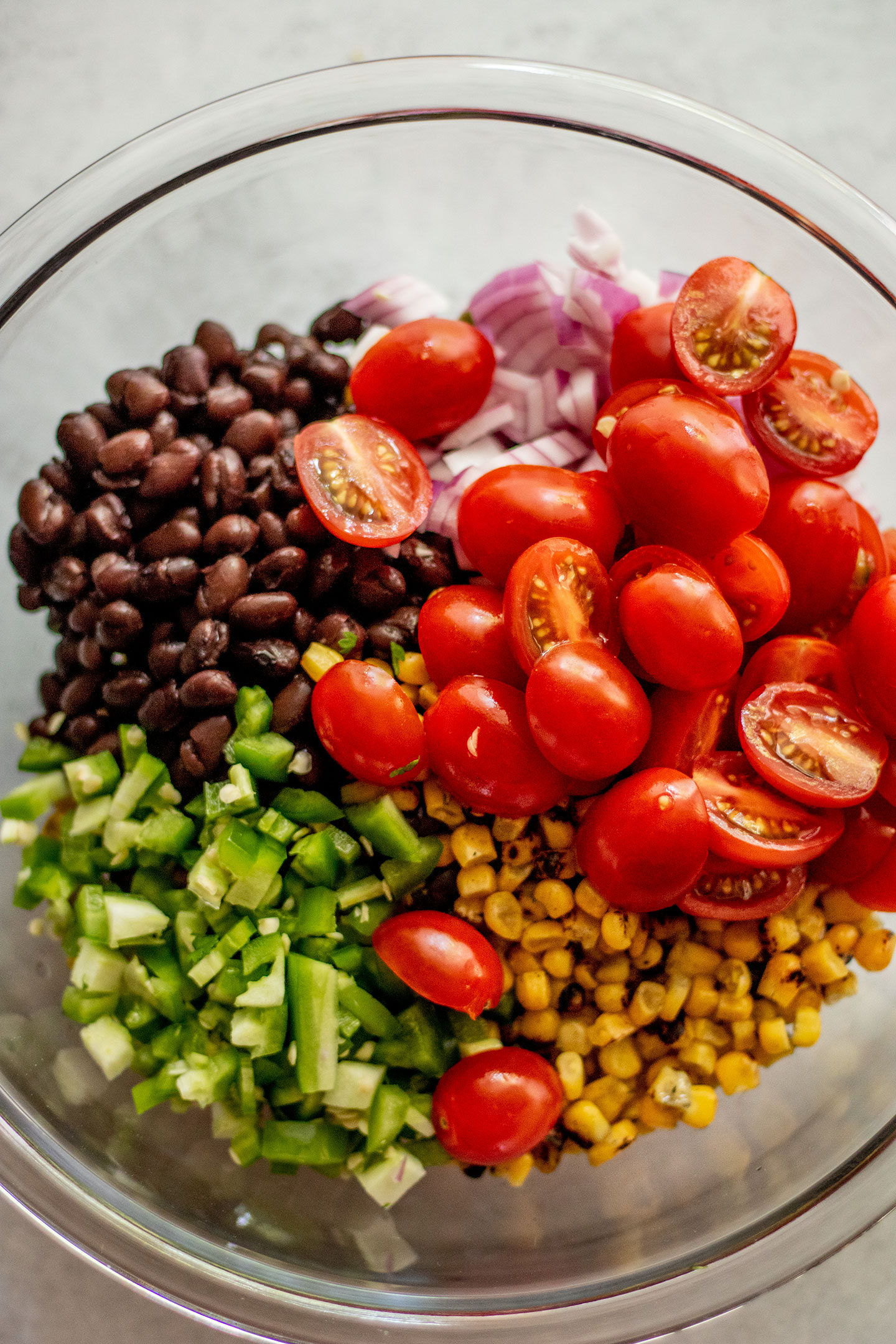 A large glass mixing bowl filled with black beans, chickpeas, cherry tomatoes, jalapeno, corn and diced onions.