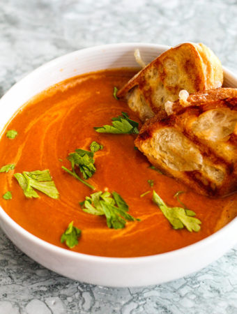 Side shot of a warm bowl of tomato soup with grilled cheese sandwiches dunked into the soup.