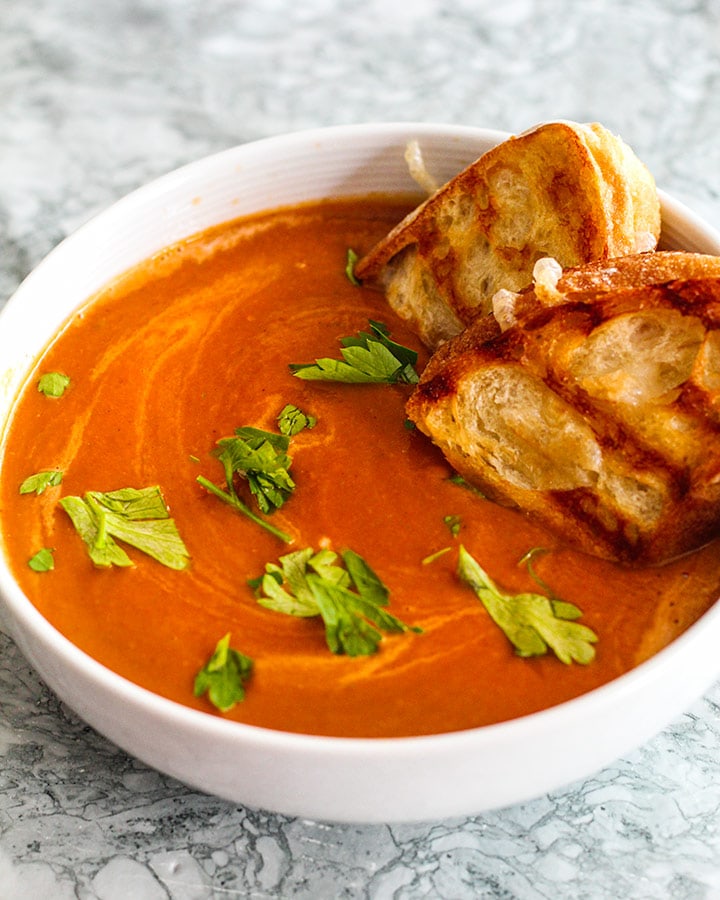 Side shot of a warm bowl of tomato soup with grilled cheese sandwiches dunked into the soup.