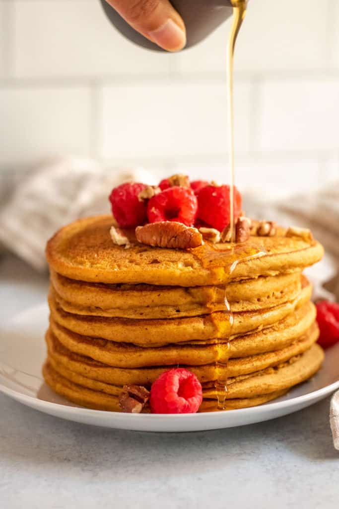 Pouring maple syrup on top of a stack of pumpkin pancakes after topping with raspberries and pecans.