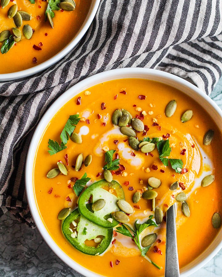 The butternut squash soup poured into two bowls and topped with jalapeno, red pepper flakes, coconut, pepitas and parsley.