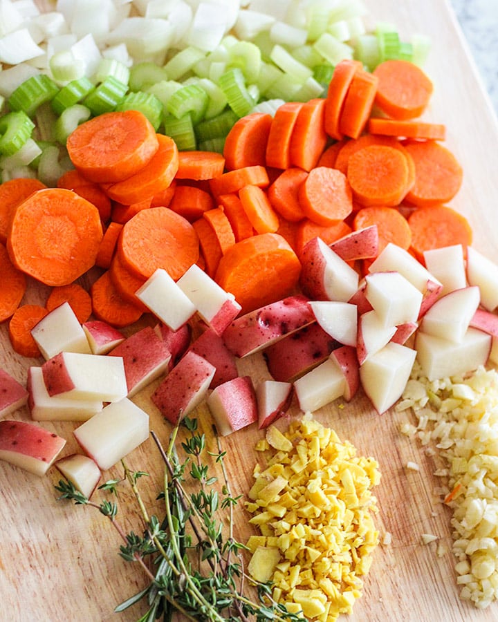 Cutting board with onions, celery, carrots, potato, fresh herbs, garlic, and ginger.