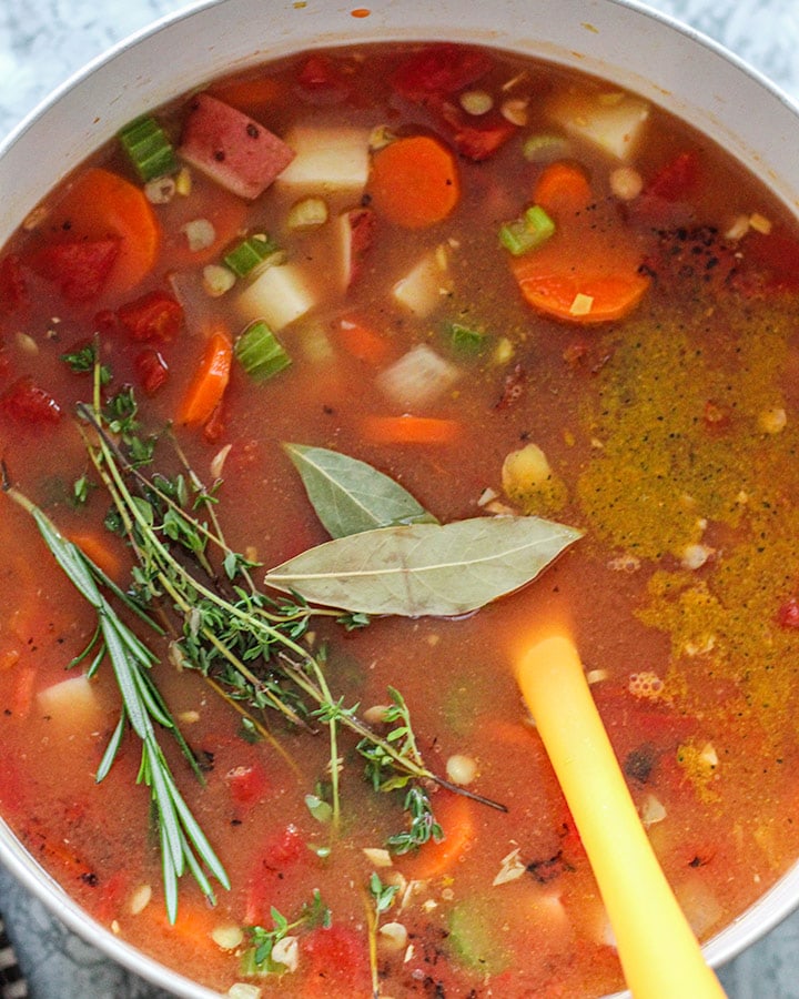 All soup ingredients as well as adobo, sazon, and fresh herbs added to the pot for cooking.