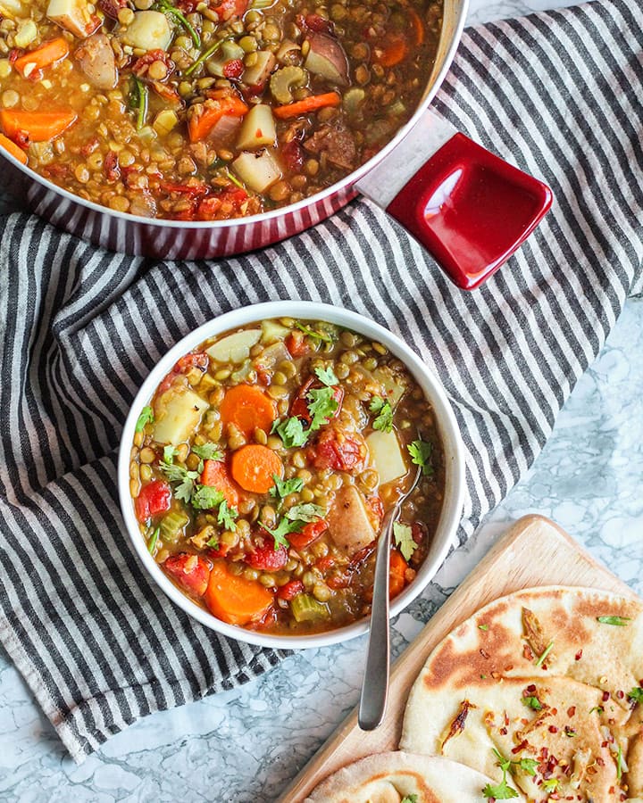 Large pot of lentil soup at the top with a bowl of soup in the center and warm pita at the bottom corner.