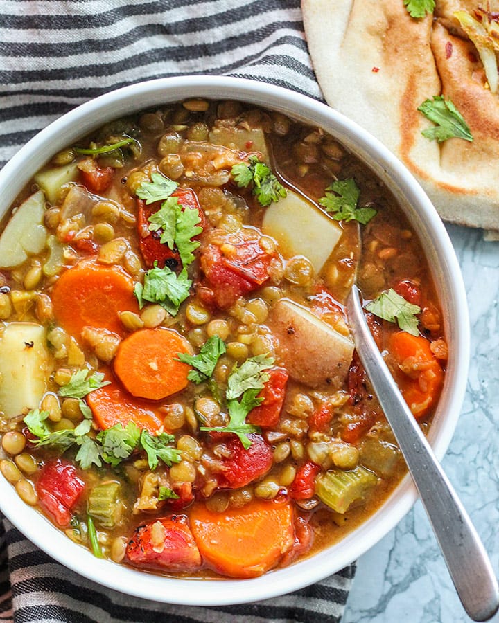 Bowl of lentil soup with a spoon pointing towards the viewer and pita in the backgroun.