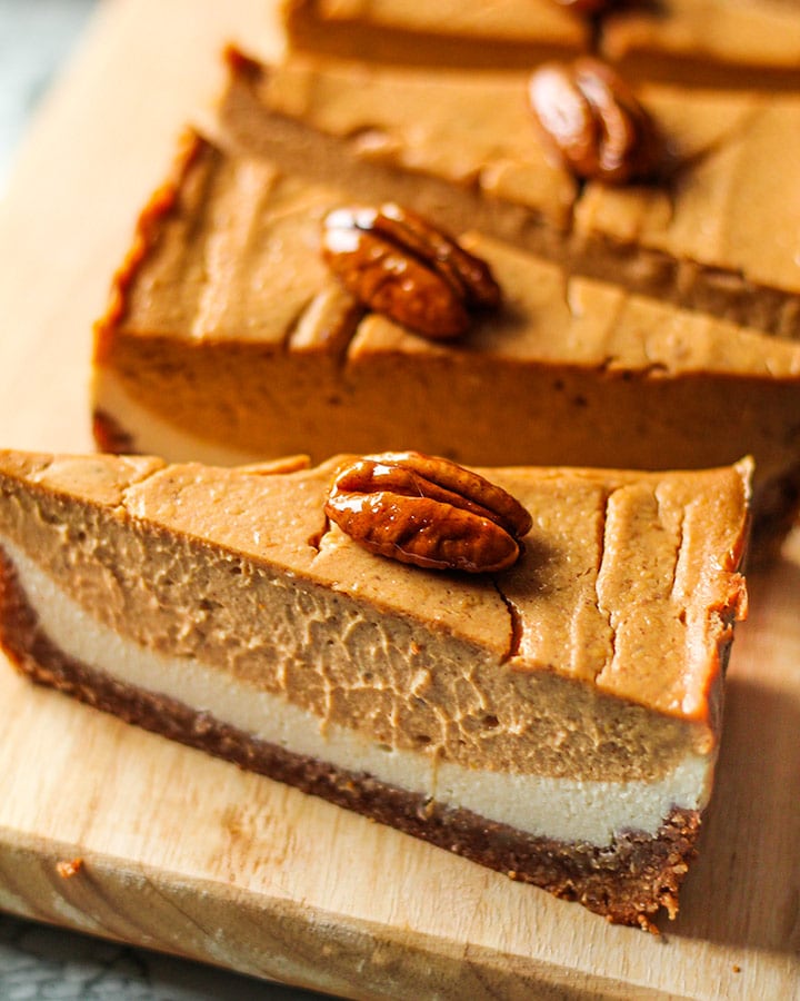 A row of double layer pumpkin cheesecake laid out on a cutting board and topped with candied pecans.