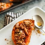 One sweet potato boat topped with crumble and pecans on top of a plate with a spoon of maple syrup and the tray of extra sweet potatoes in the background.