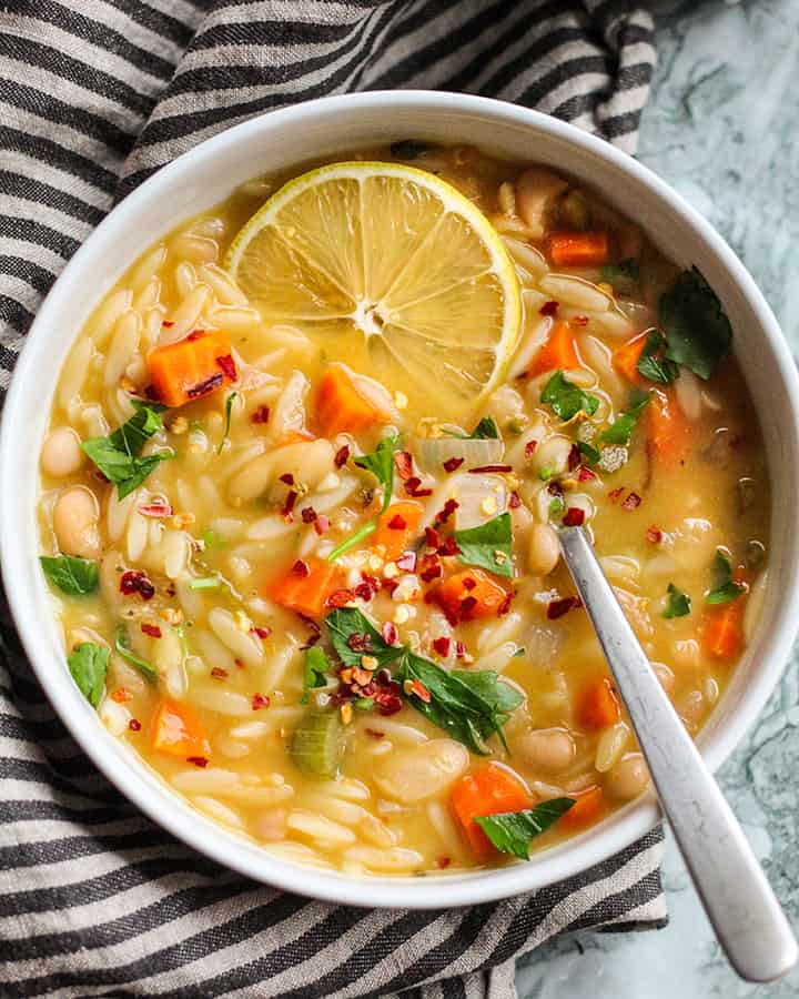 A bowl of orzo soup with a slice of lemon on top with a sprinkle of parsley and red pepper flakes and a spoon to the side.