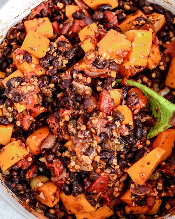 Mixing the butternut squash, tomatoes, black beans and red lentils together with the spices that cooked in the pot with a large green spoon.
