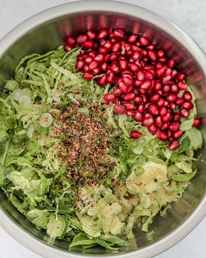 A mixing bowl with added shaved brussel sprouts, scallions, herbs and spices, and pomegranate.