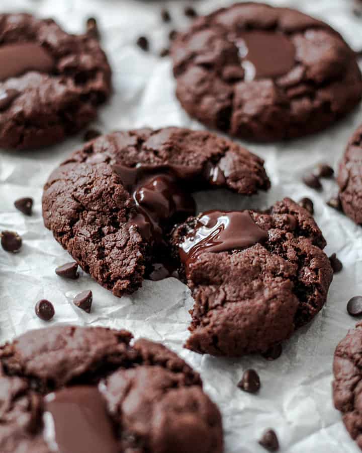 Side view of the tray of cookies with a scatter of chocolate chips around them. Middle cookie is pulled apart to show melted chocolate.