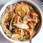 Bowl of spiced oatmeal topped with sautéed pears, pecans, peanut butter, maple syrup, and pepitas.
