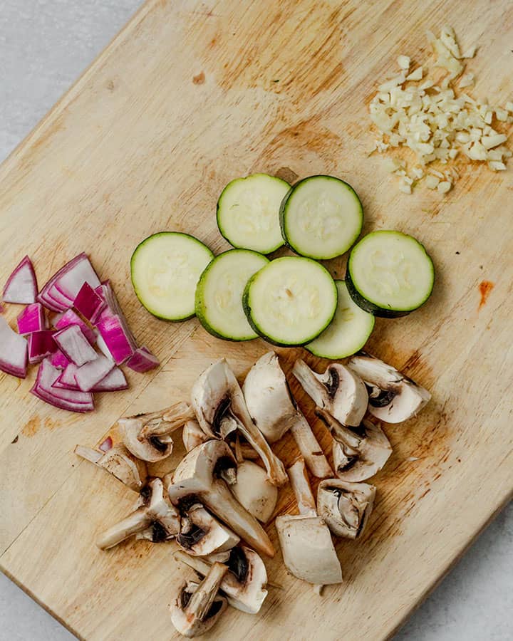 Cutting board with freshly chopped vegetables including red onion, mushrooms, zucchini and garlic. These vegetables are prepared for sautéing in the next step of this recipe.