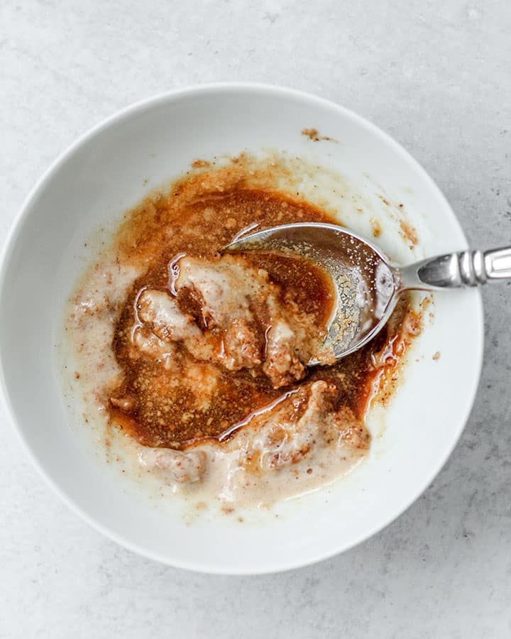 In a white bowl you see a mixture of almond butter, miso paste, soy sauce, vinegar, spices and sweetener being mixed together with a silver spoon. This almond miso sauce will be poured over the base ingredients of the bowl at the end.