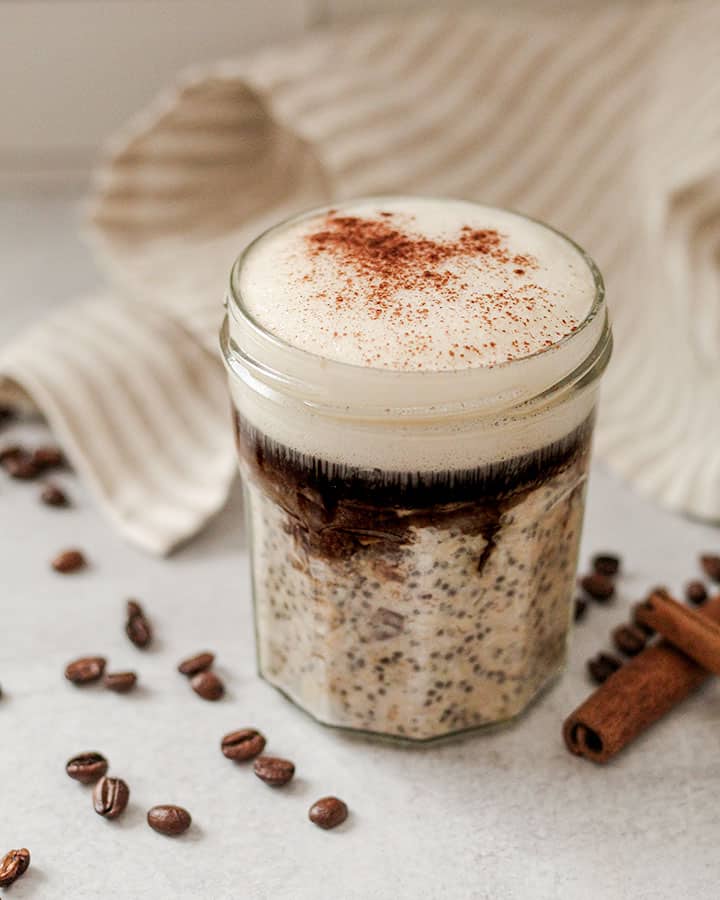 Jar of overnight oats with towel in the background and coffee beans and cinnamon sticks. 