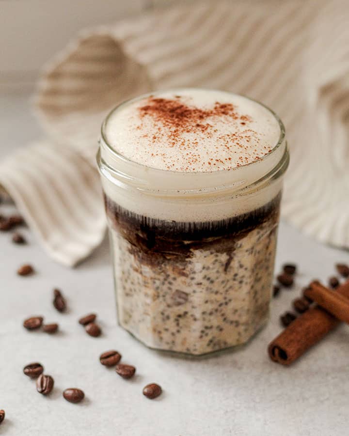 Overnight oats topped with fresh brewed coffee, foamed plant milk and sprinkled with a pinch of cinnamon on top. Napkin in the background with whole coffee beans and cinnamon sticks scattered around the jar.