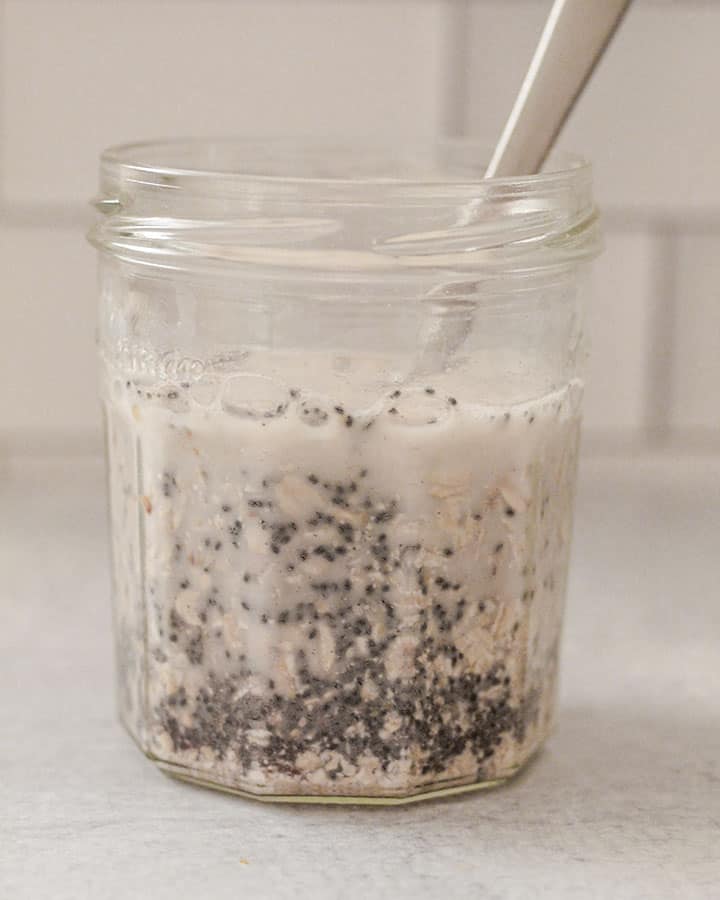 Jar of overnight oats being set up with plant milk and mixed together with a silver matte spoon.