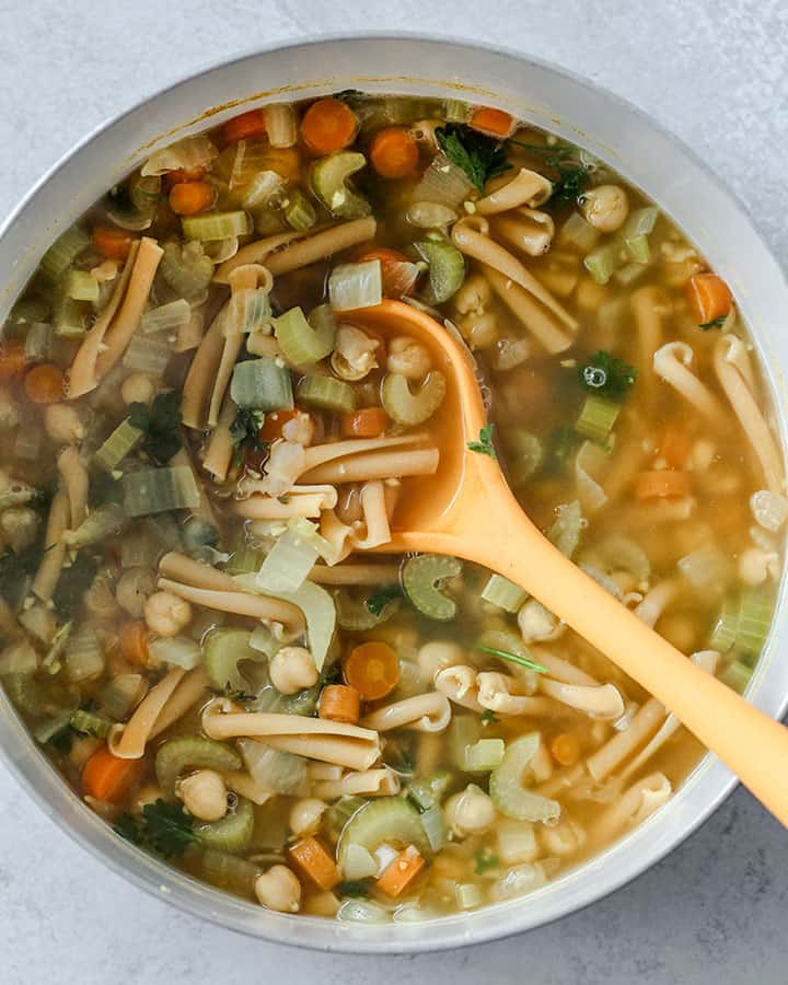 Large pot of soup being stirred with a yellow ladle, which is scooping up some noodles, vegetables and chickpeas.