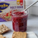 Jar of raspberry jam with a serving knife dipped inside. Cutting board in front has a crispbread spread with peanut butter. The background has a container of Wasa crispbreads in the background.