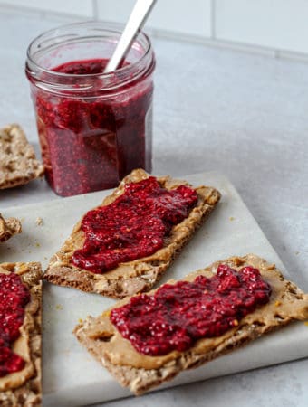 Crispbreads spread with peanut butter and raspberry jam on a white marbled cutting board with a jar of jam in the background with a spreading knife.