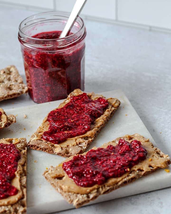 Crispbreads spread with peanut butter and raspberry jam on a white marbled cutting board with a jar of jam in the background with a spreading knife.