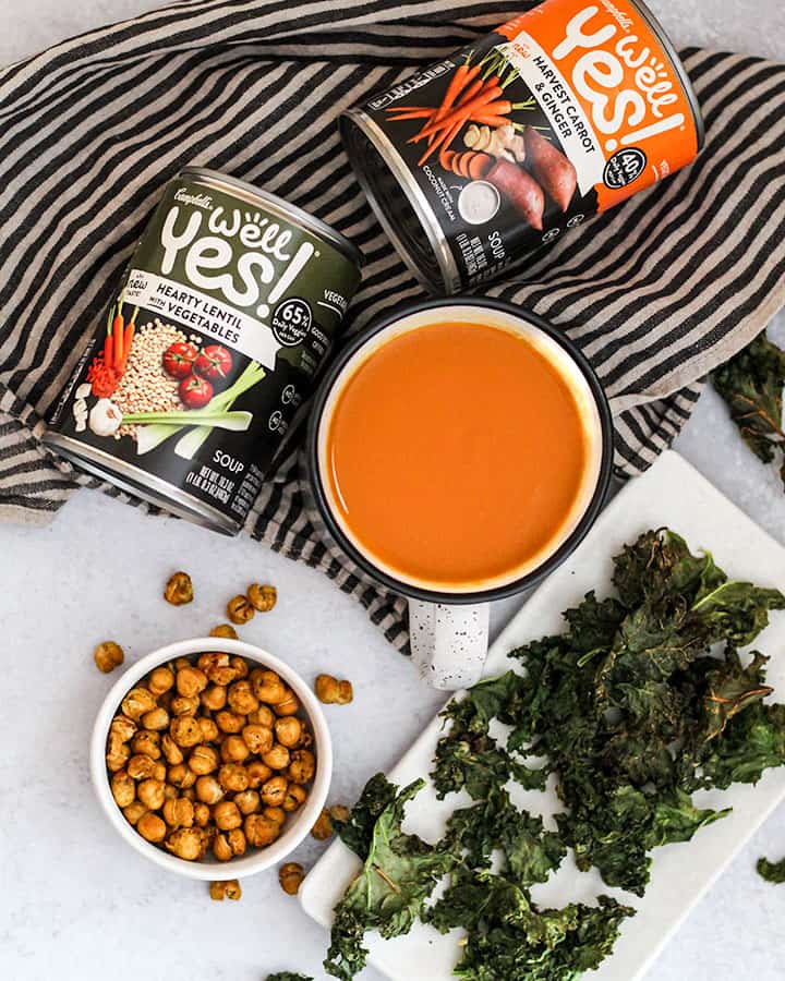 Cans of soup at the top with roasted chickpeas and roasted kale plated at the bottom of the picture and a large mug of harvest carrot and ginger soup in a wide mouth mug. This paints a picture of how important it is to use realistic vegan shopping tips to make being and staying vegan easier.