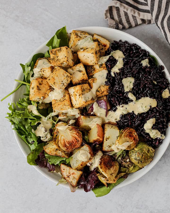 White bowl that has baked tofu, vegetables on top of greens and paired with black rice and topped with ranch. A stripped napkin can be seen peaking in the background.