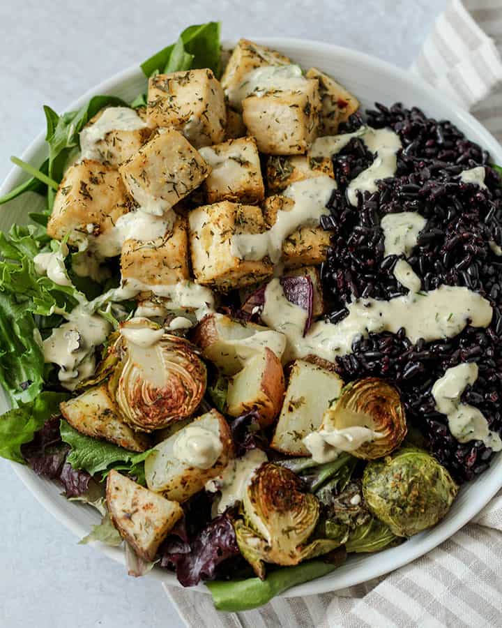 Close up of sheet pan bowl that includes greens, sheet pan vegetables, baked tofu and black rice topped with ranch.
