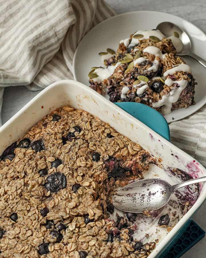 Viewing the baked oatmeal dish in front with a plate behind it housing a scoop of the baked oats topped with plant yogurt.