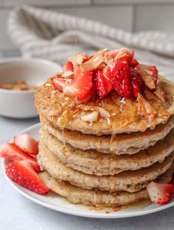 Stack of pancakes with syrup dripping from the sides and with toasted coconut flakes in the back for topping.