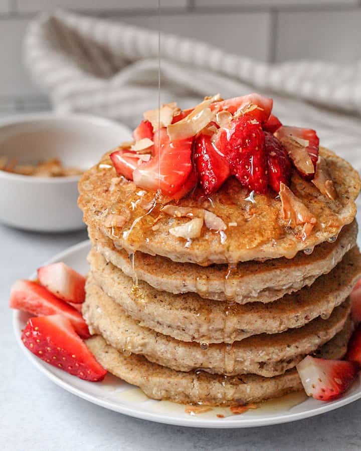 Stack of pancakes with syrup dripping from the sides and with toasted coconut flakes in the back for topping.