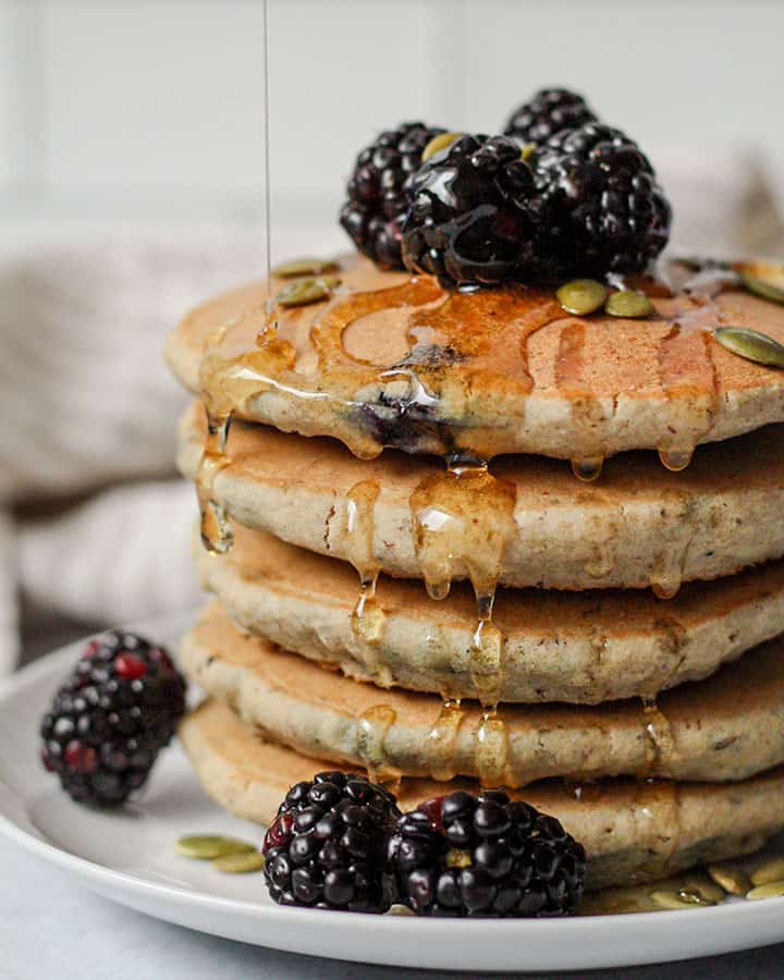 A stack of blueberry pancakes stacked and topped with pepitas and fresh blackberries with a slow drizzle of syrup being poured overtop.