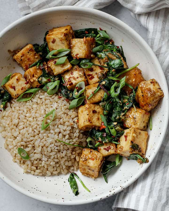 Top view of the a bowl of brown rice served with garlic "honey" tofu on the side with wilted spinach and sliced spring onion.