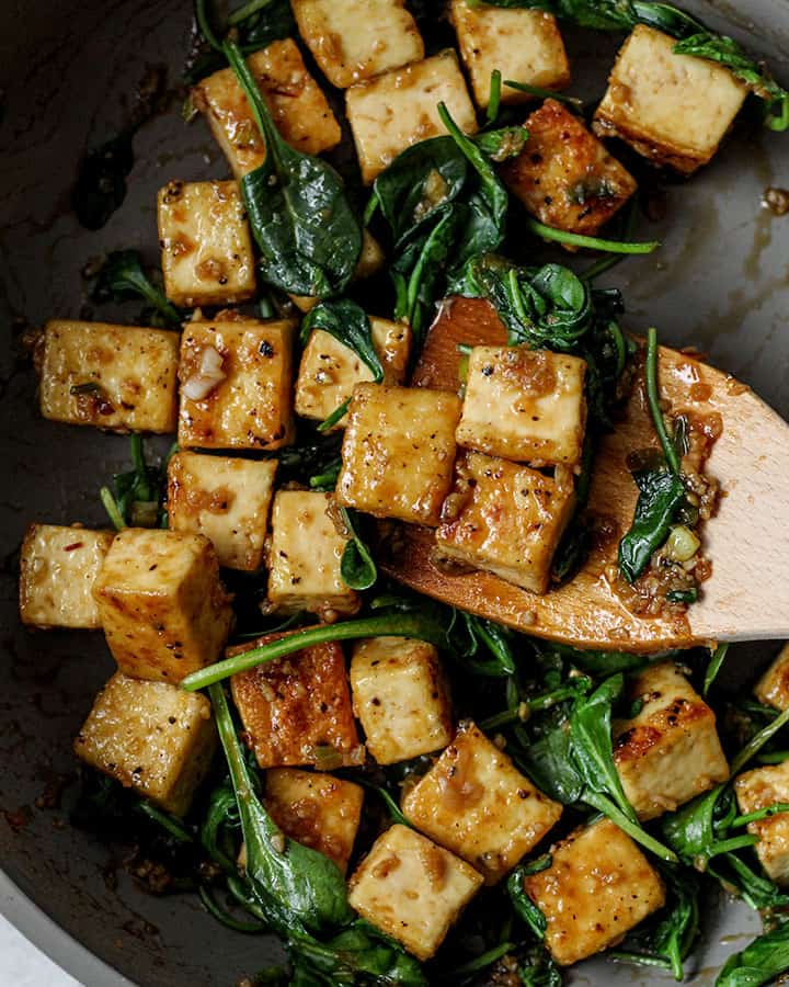 Cooked tofu coated with garlic "honey" while being stirred together with wilted spinach in a pan using a wooden spoon.