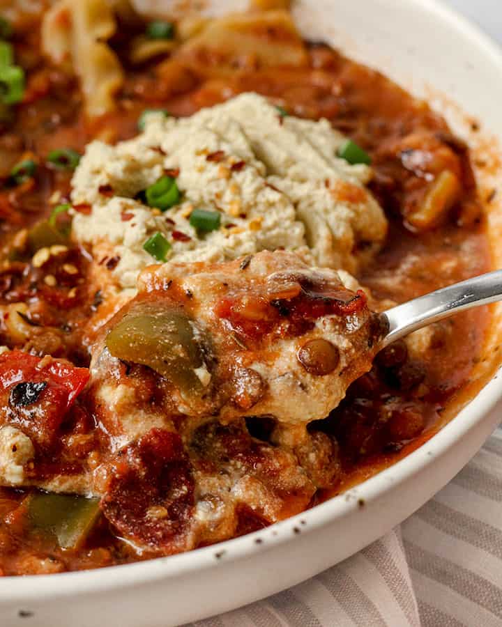 Side view close up of the lasagna soup with a spoon lifting a portion of the soup up along with a generous amount of vegan ricotta.