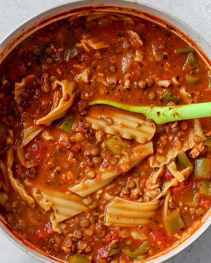 Fully cooked lasagna soup, with lasagna noodles being stirred into the soup with a ladle. 